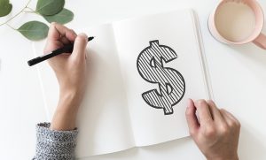 Image is a hand drawing a dollar sign in a notebook that is sitting next to a cup of coffee. Image is a hand drawing a dollar sign in a notebook that is sitting next to a cup of coffee.